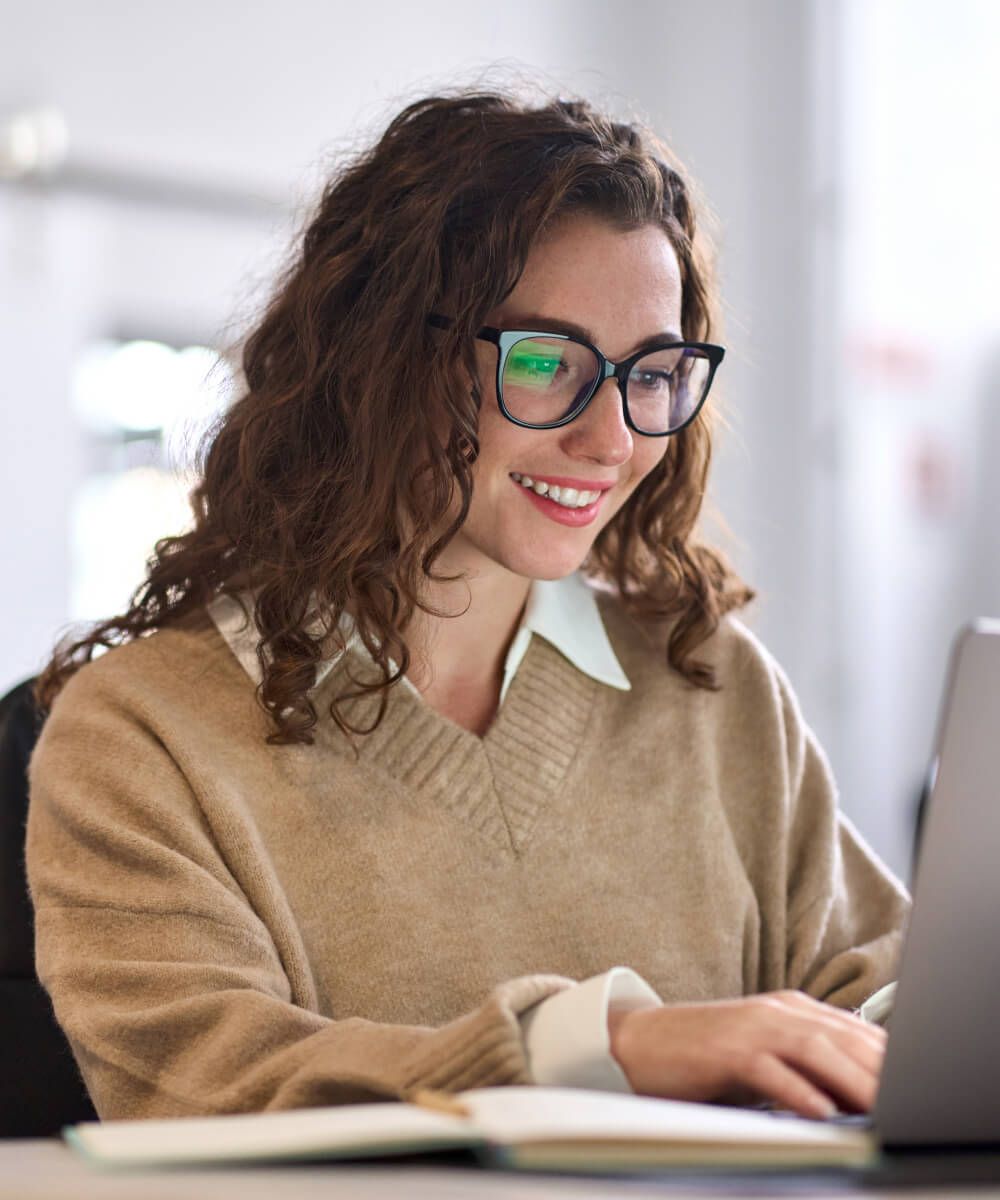 Smiling woman with glasses working on laptop at a desk in a bright modern workspace - Neutrinos